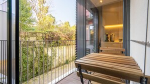 A balcony with a wooden bench, a glass door leading to a room with a coffee machine and a plant, and a view of a garden with trees and a stone wall.