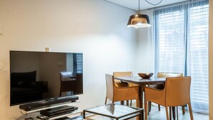 A modern living room with a flat-screen TV, a glass coffee table, and a dining area with four chairs.