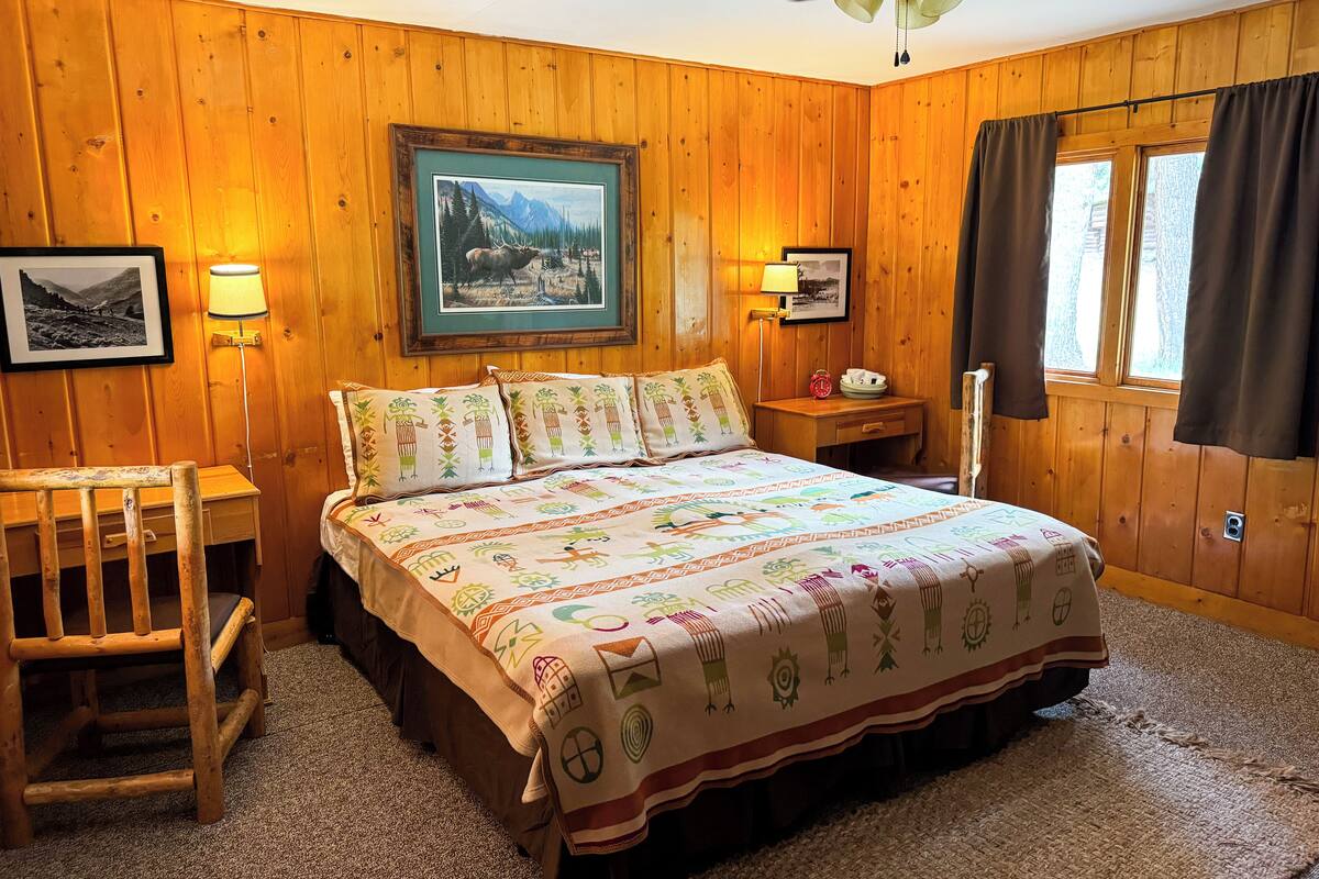 A wooden-paneled room with a bed, two bedside lamps, a framed picture on the wall, and a window with curtains.