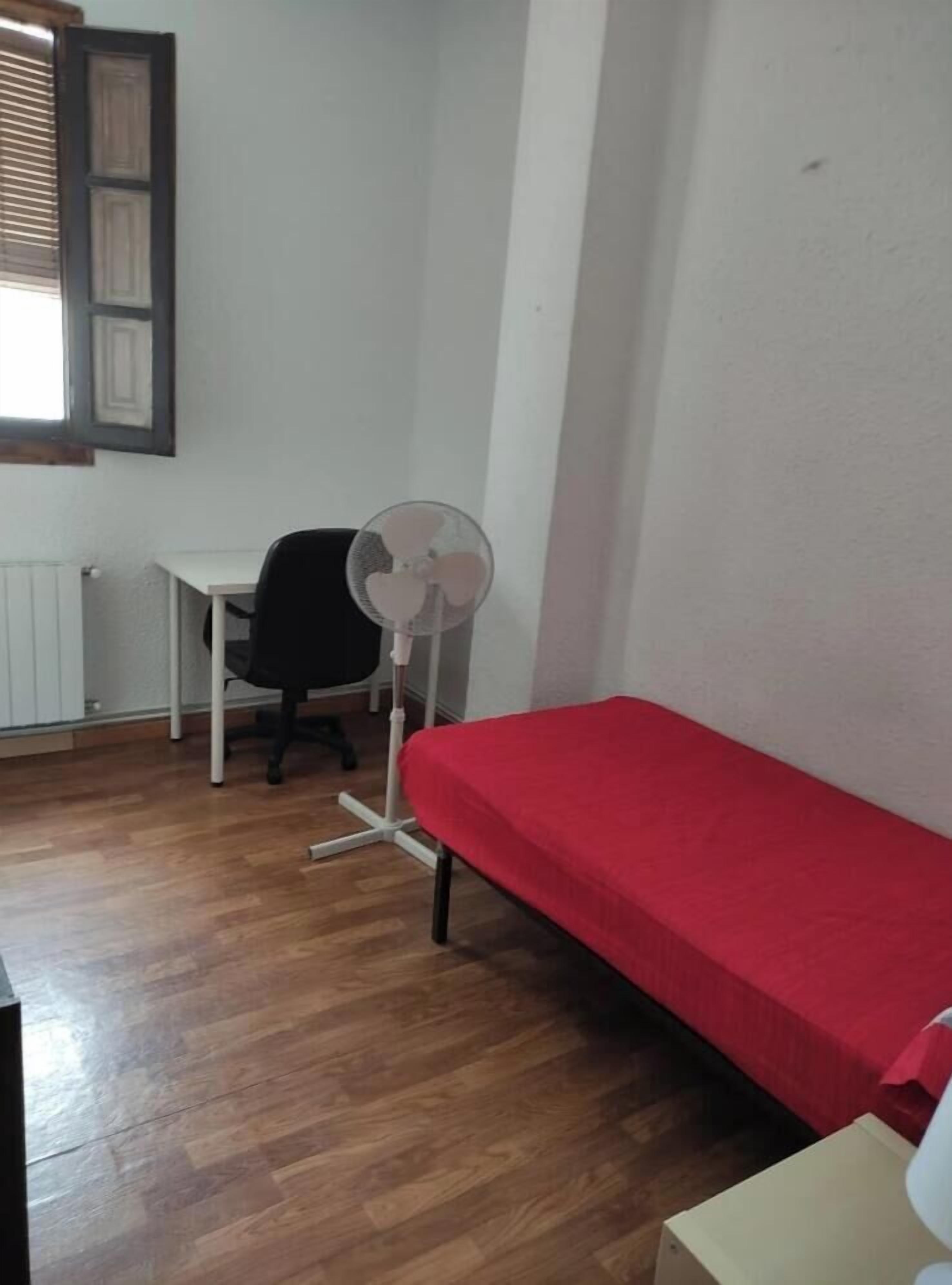 A small room with a single bed, a desk, a chair, and a standing fan.