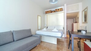Ett modernt hotellrum med en grå soffa, en snyggt bäddad säng, ett litet bord med en mugg och en garderob med resväskor.