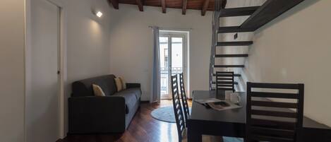 Ein kompakter Wohnraum mit einem dunklen Sofa, einem Esstisch mit Stühlen und einer Treppe.