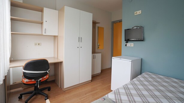 Ein Schlafzimmer mit einem Bett, einem Schreibtisch mit Stuhl, einem Kleiderschrank, einem Fernseher und einem kleinen weißen Schrank.
