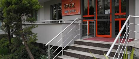 A hotel entrance with a red and white facade, a staircase leading to the entrance, and a small garden area with plants.