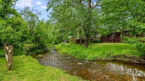 Ein Fluss fließt durch eine üppige, grüne Landschaft mit einem hölzernen Zaun und einem Gebäude mit Terrasse im Hintergrund.