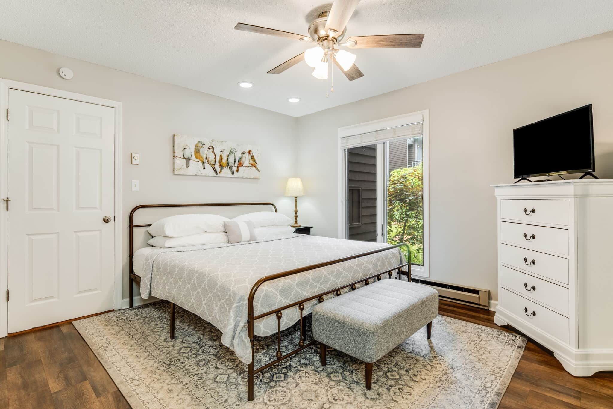 A bedroom with a bed, a ceiling fan, a TV, a dresser, and a window.