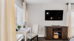 Un espacio habitable compacto con un escritorio blanco, una silla, un televisor montado en la pared y un armario de madera con almacenamiento.