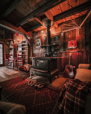 A rustic room with a large wood-burning stove, wooden walls, a guitar, and a plaid blanket on the floor. - Enjoy this authentic and cozy VT Barn with Brand New Hot Tub (Londonderry)