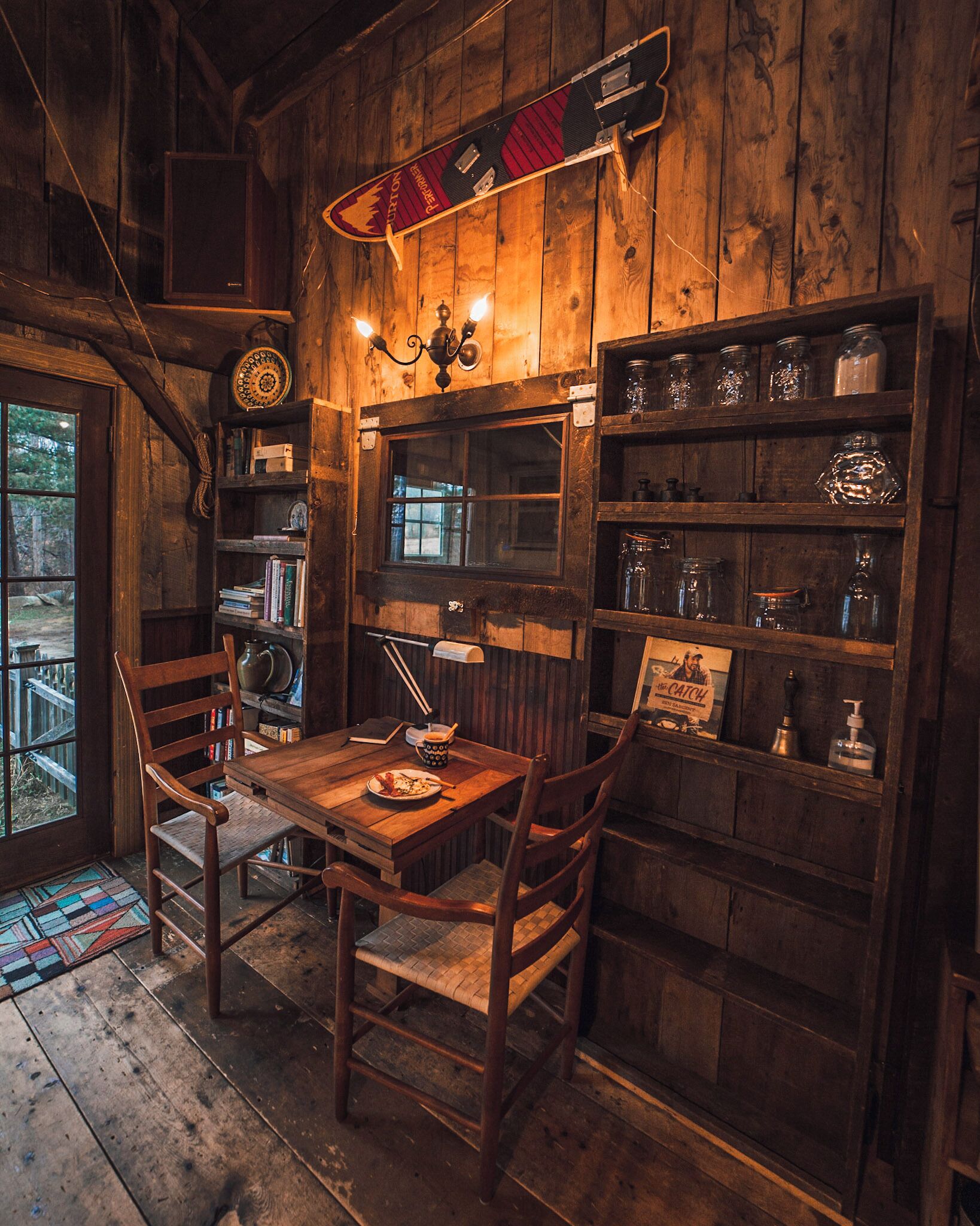 Un intérieur rustique en bois, avec une table à manger, des chaises, une bibliothèque et une planche de surf.