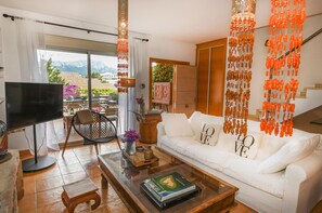 Un salon avec un canapé blanc, une table basse, un téléviseur et une vue sur les montagnes, visible à travers la porte vitrée.