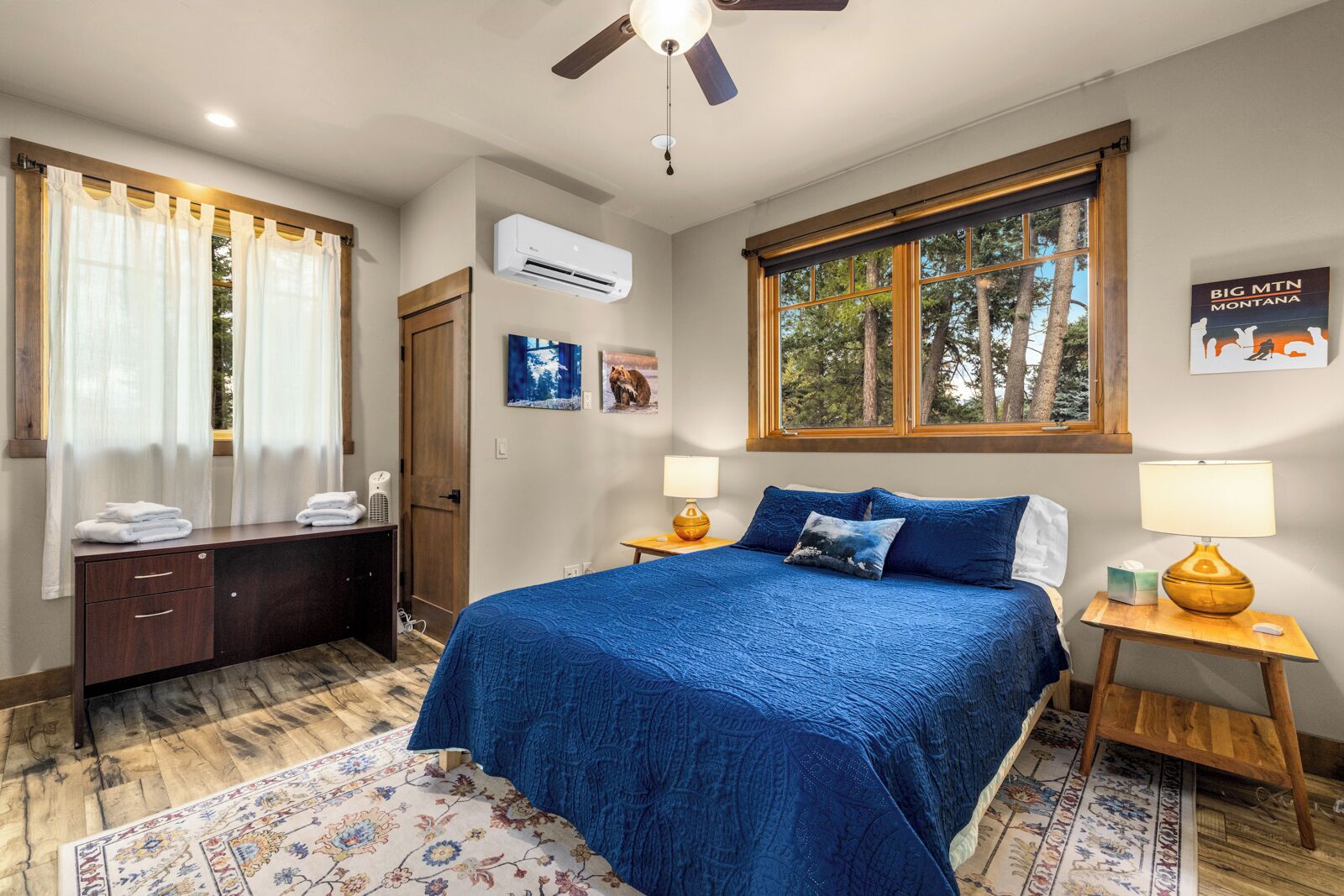 A bedroom with a large bed, two bedside tables with lamps, a wooden dresser, a window with a view of trees, and an air conditioning unit.