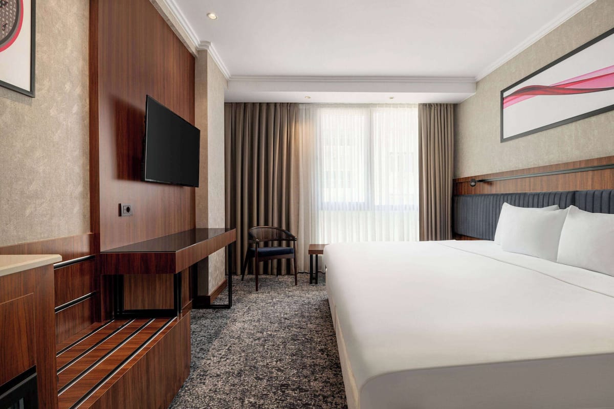 Une chambre d’hôtel dotée d’un grand lit, d’un téléviseur à écran plat fixé au mur, d’un bureau avec une chaise et d’une fenêtre avec des rideaux.