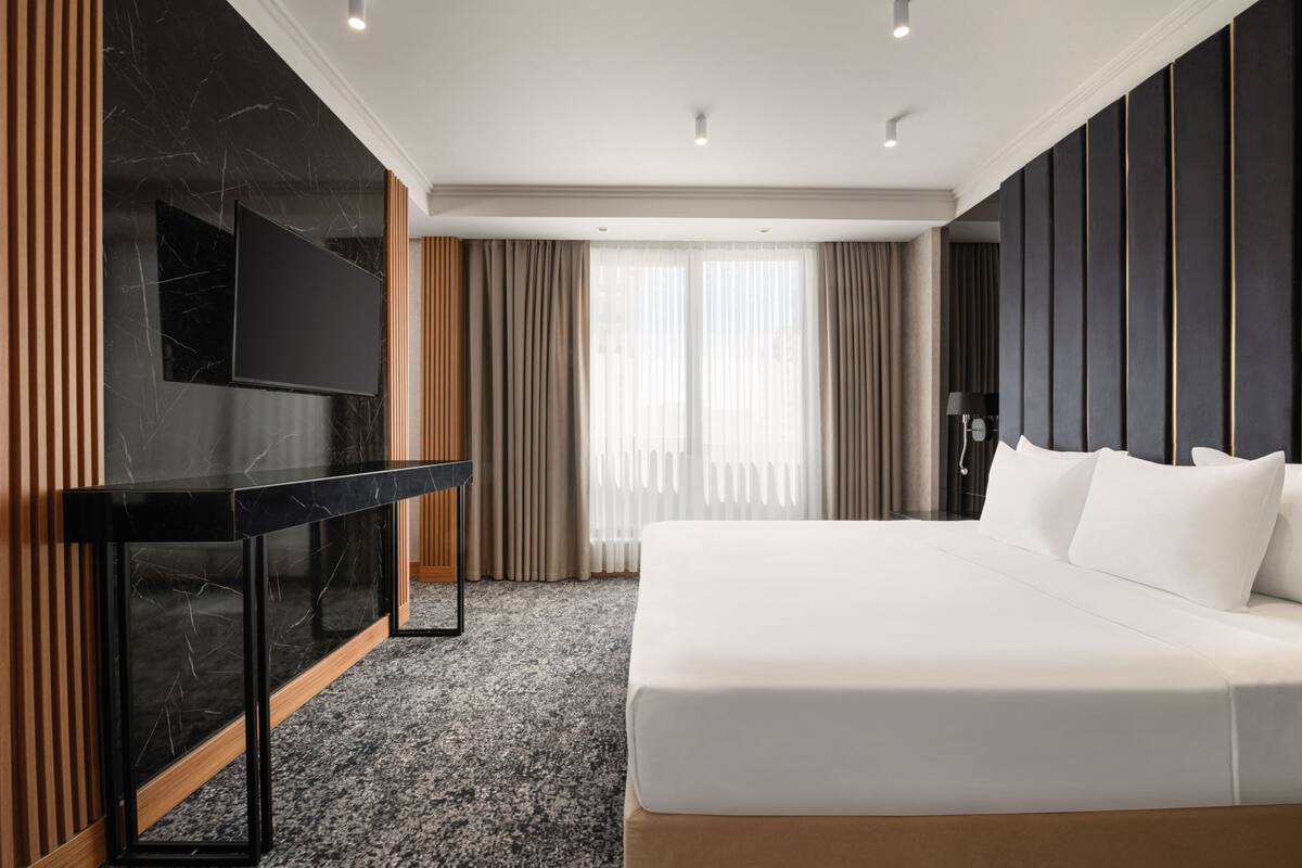 Une chambre d’hôtel moderne dotée d’un grand lit, d’un téléviseur à écran plat fixé sur une console en marbre et d’une grande fenêtre avec des rideaux.