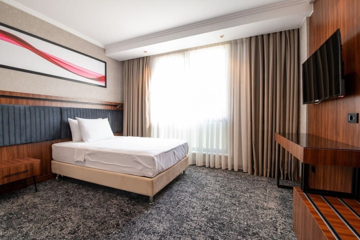 Une chambre d’hôtel avec un lit, une télévision, un bureau en bois et une grande fenêtre avec des rideaux.