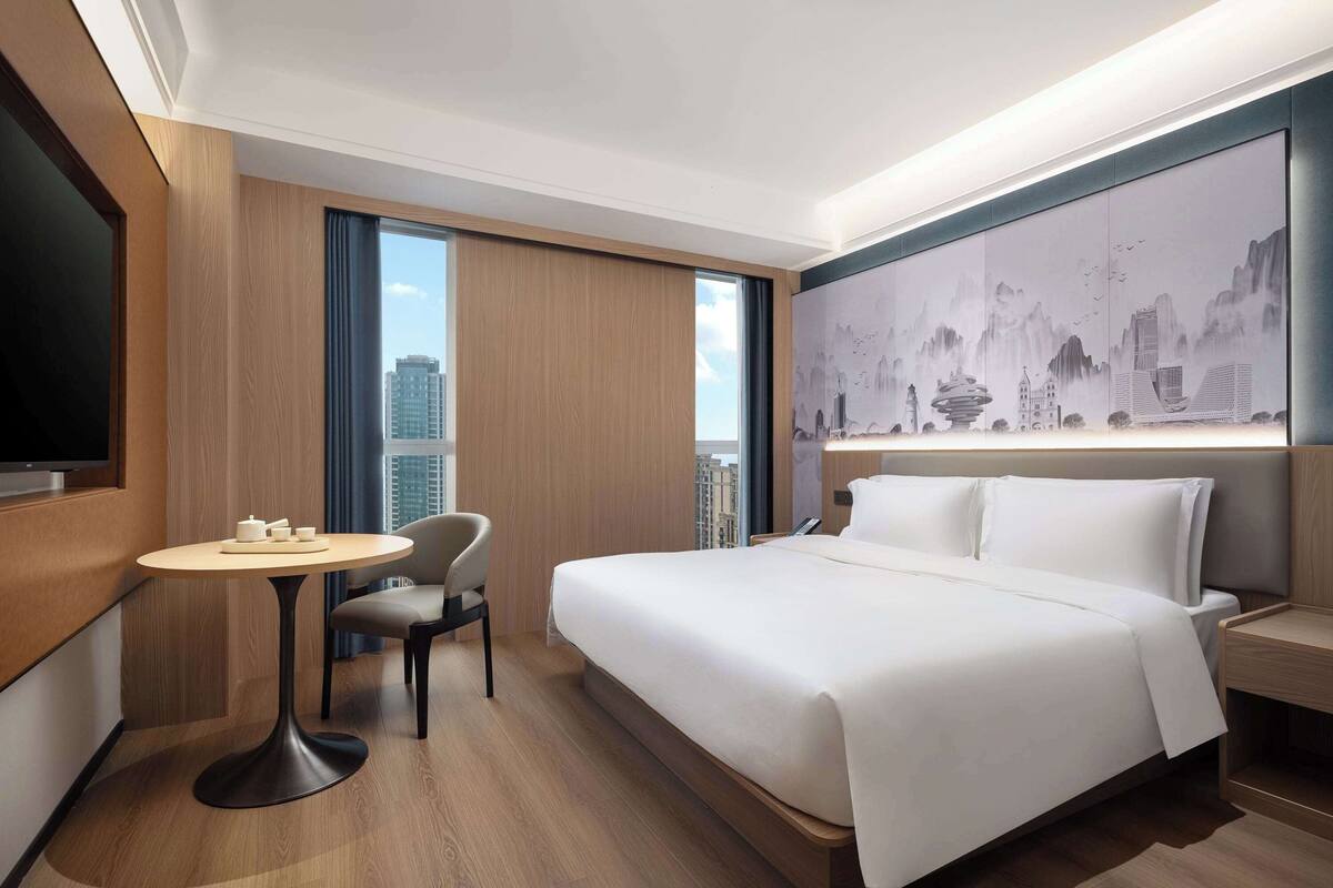 Ein modernes Hotelzimmer mit einem großen Bett, einem runden Tisch, einem Stuhl und einem Stadtbild an der Wand.