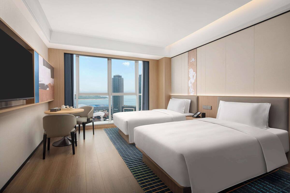 Ein Hotelzimmer mit zwei Betten, einem Schreibtisch und einem Stuhl. Ein großes Fenster bietet Blick auf die Stadt.