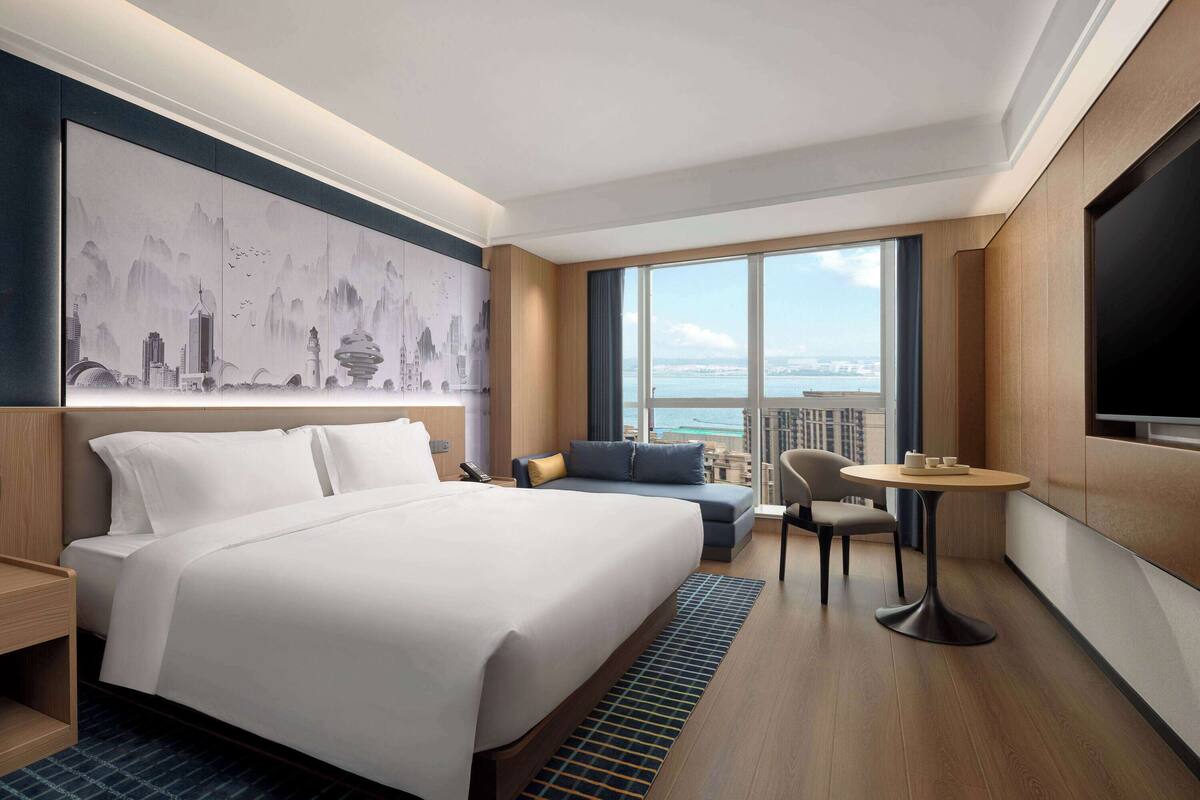 Ein Hotelzimmer mit einem großen Bett, einer Couch, einem kleinen Tisch und einem Fernseher. Das Zimmer verfügt über ein großes Fenster mit Blick auf die Stadt.