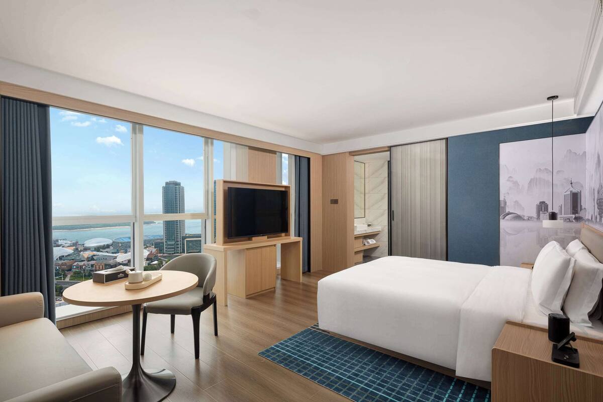Ein modernes Hotelzimmer mit einem großen Bett, einem Schreibtisch mit Fernseher, einer Sitzecke mit Tisch und Blick auf die Stadt.