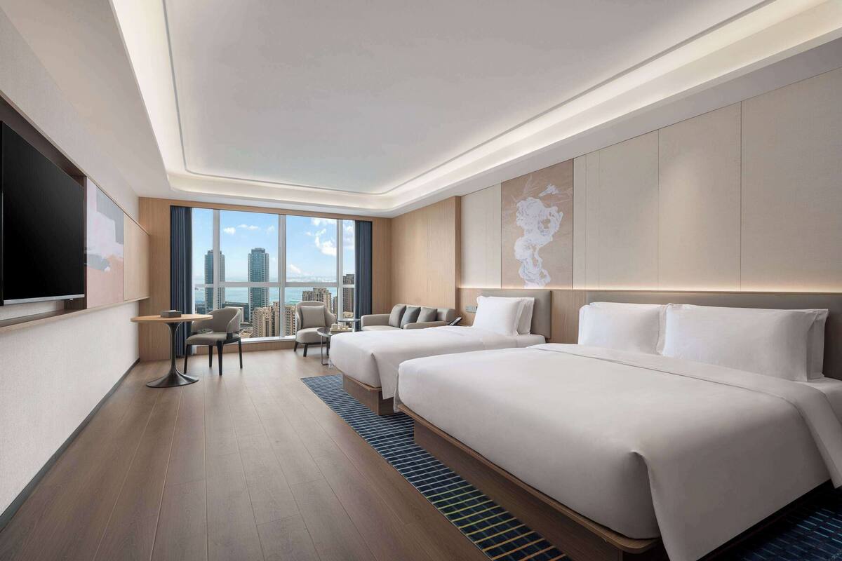 Ein modernes Hotelzimmer mit zwei Betten, einem Flachbildfernseher, einem kleinen Tisch und Blick auf die Stadtlandschaft.
