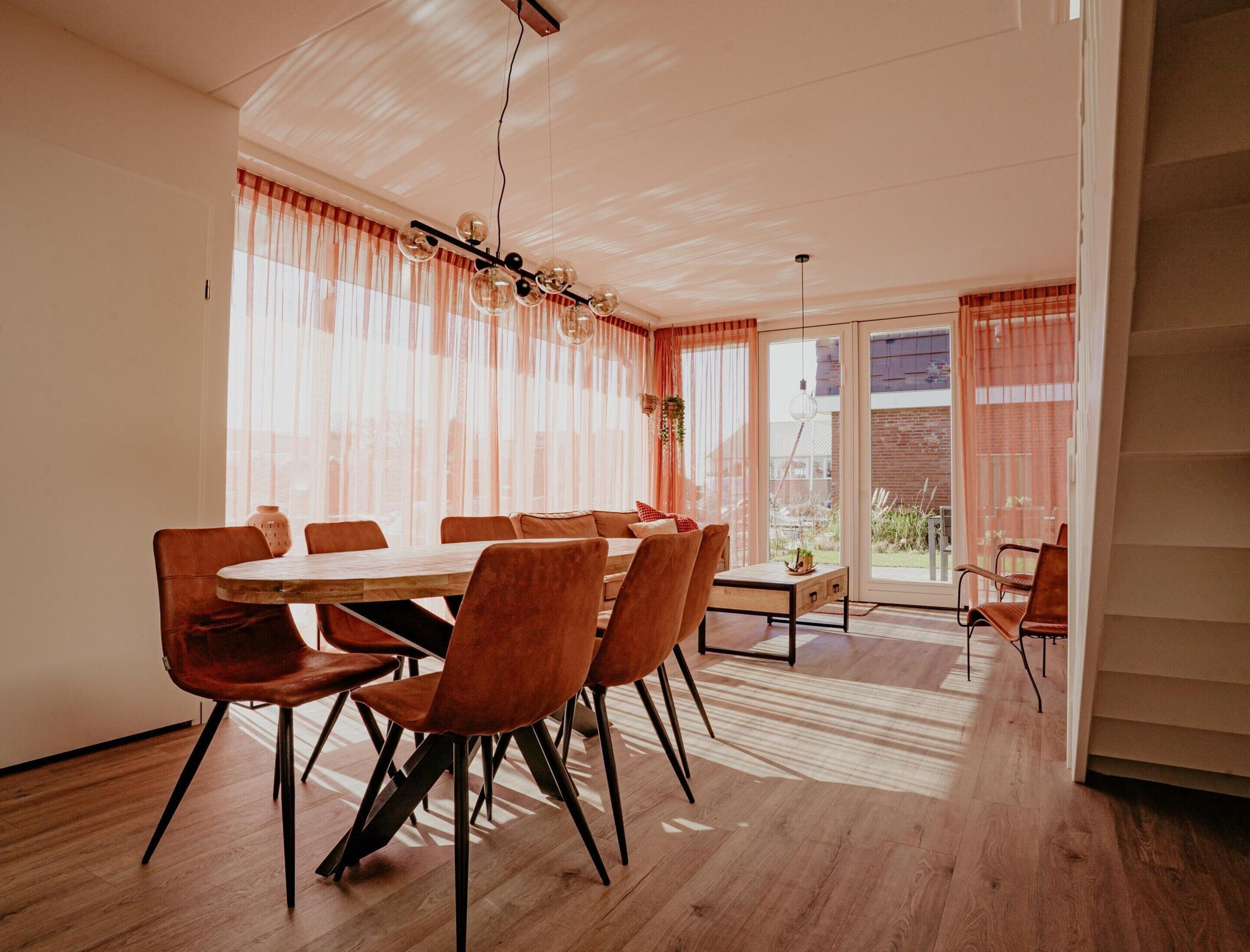 A dining area with a wooden table and chairs, large windows with sheer curtains, and a view of the outside.