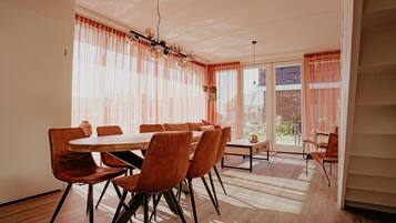 A dining area with a wooden table and chairs, large windows with sheer curtains, and a view of the outside.