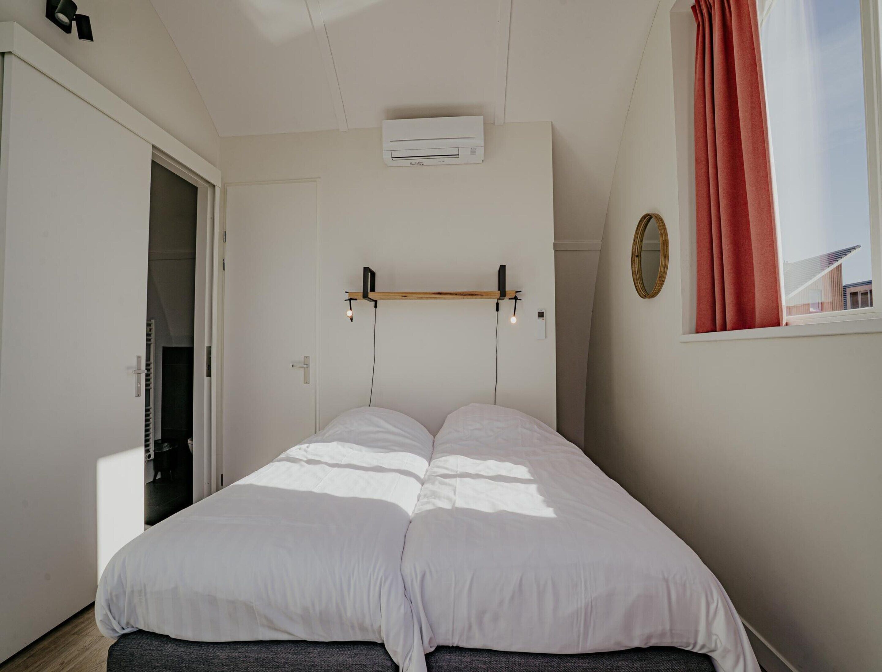 Ein Schlafzimmer mit einem Bett, einer Klimaanlage, einem Fenster mit Vorhang und einem Spiegel an der Wand.