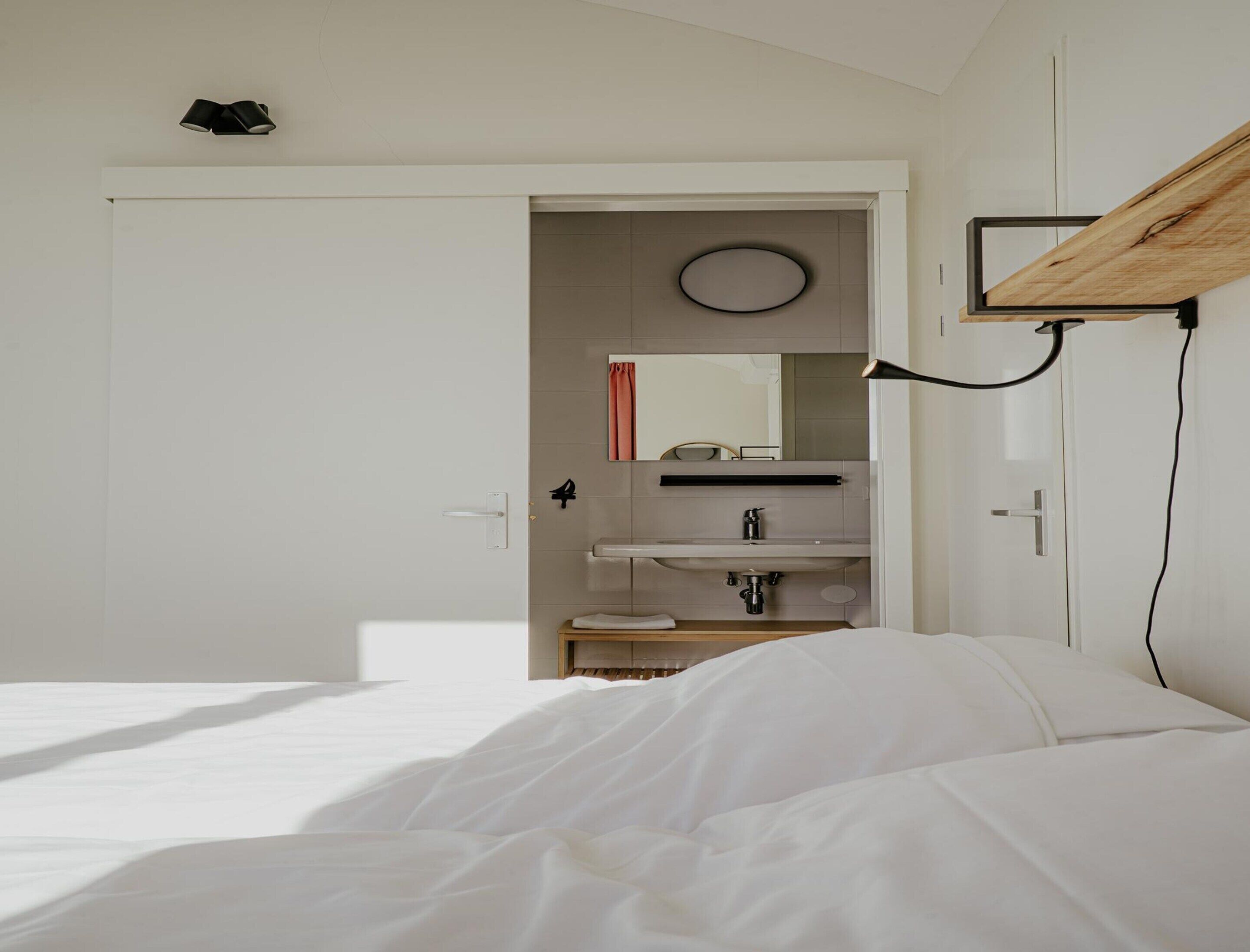 A bedroom with a bed, a white wall, a door, a mirror, a sink, and a towel rack.