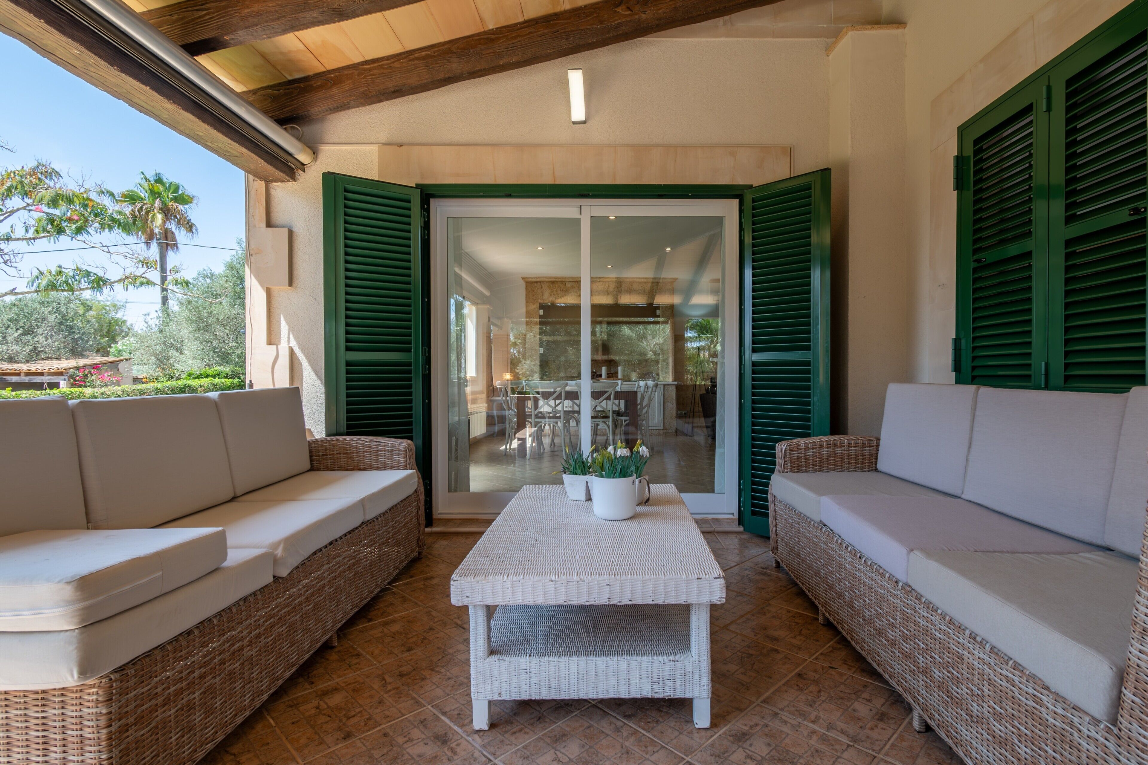 A covered outdoor seating area with wicker furniture and a white table, a glass door with green shutters, and a view of the interior.