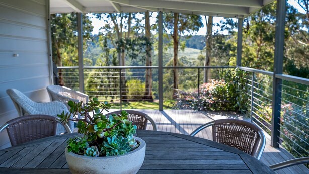 Un área de estar al aire libre con techo, una mesa y sillas, una planta en maceta, y vistas a un jardín y árboles.