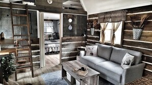 A rustic cabin interior with a grey sofa, wooden ladder, and a dining table. - Southern Blooms! Porch Swinging, Pet-Friendly, Tiny Home near Kentucky Lake! (Springville)