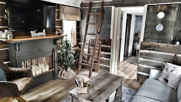 A rustic interior with a wooden TV stand labeled "Unwind," a ladder, a Christmas tree, and a cozy seating area. - Southern Blooms! Porch Swinging, Pet-Friendly, Tiny Home near Kentucky Lake! (Springville)