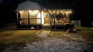 A small, single-story cabin with a porch, a welcome sign, and a flag. - Southern Blooms! Porch Swinging, Pet-Friendly, Tiny Home near Kentucky Lake! (Springville)