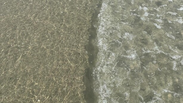 Ein Strand mit klarem, flachen Wasser, einem Sandufer und einigen Spaziergängern.