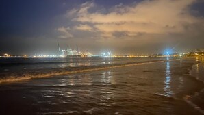 Ein Strand mit Wellen, eine entfernte Industrieanlage und ein bewölkter Himmel.