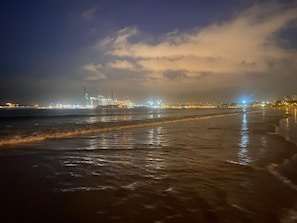 Una playa con olas, una zona industrial a lo lejos y un cielo nublado.