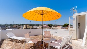 A rooftop terrace with a yellow umbrella, white lounge chairs, and a table.