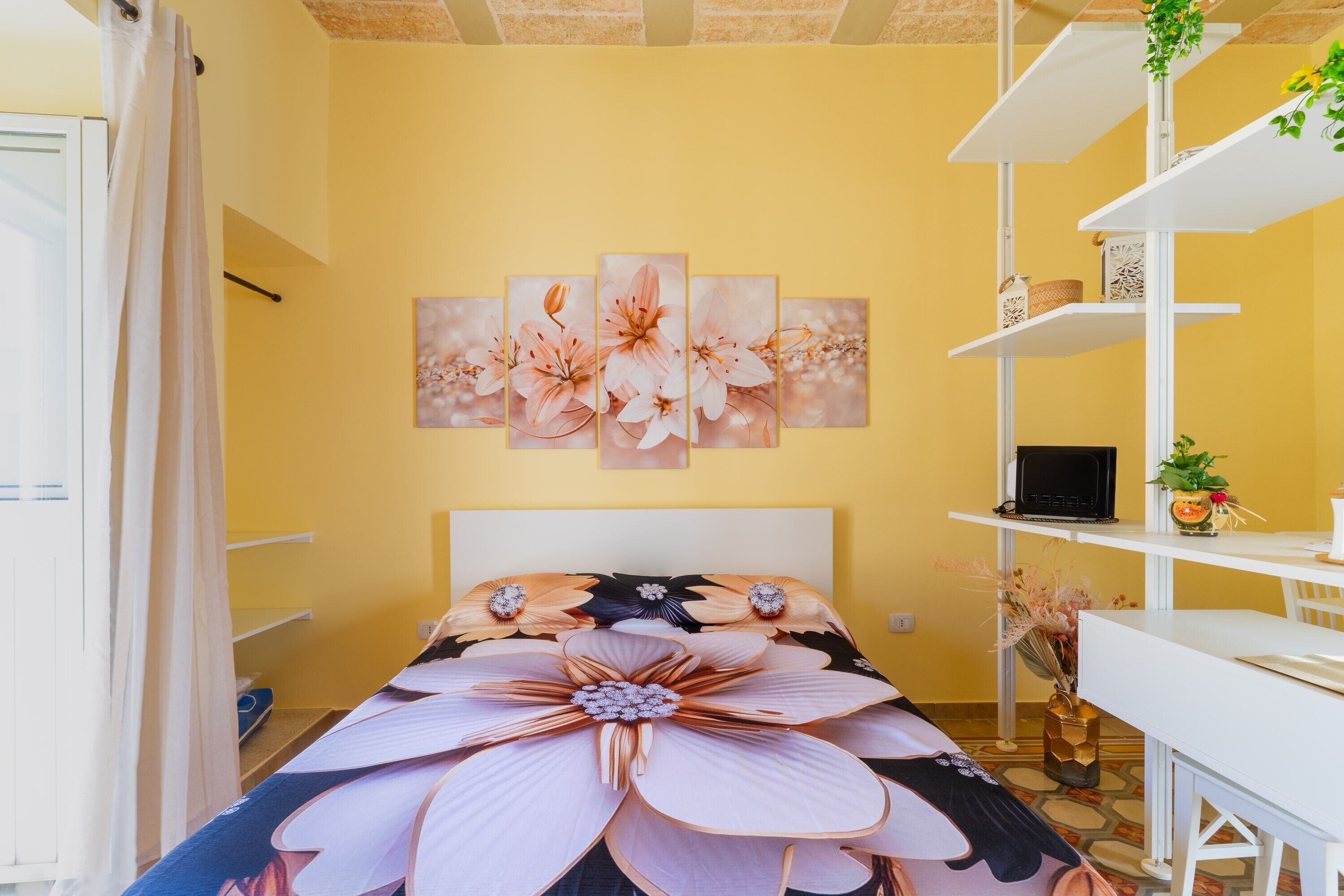 A bedroom with a bed, a desk, a chair, a shelf with a laptop, and a wall with a floral painting.