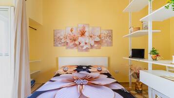 Ein Schlafzimmer mit einem Bett, einem Schreibtisch, einem Stuhl, einem Regal mit einem Laptop und einer Wand mit einem Blumenbild.