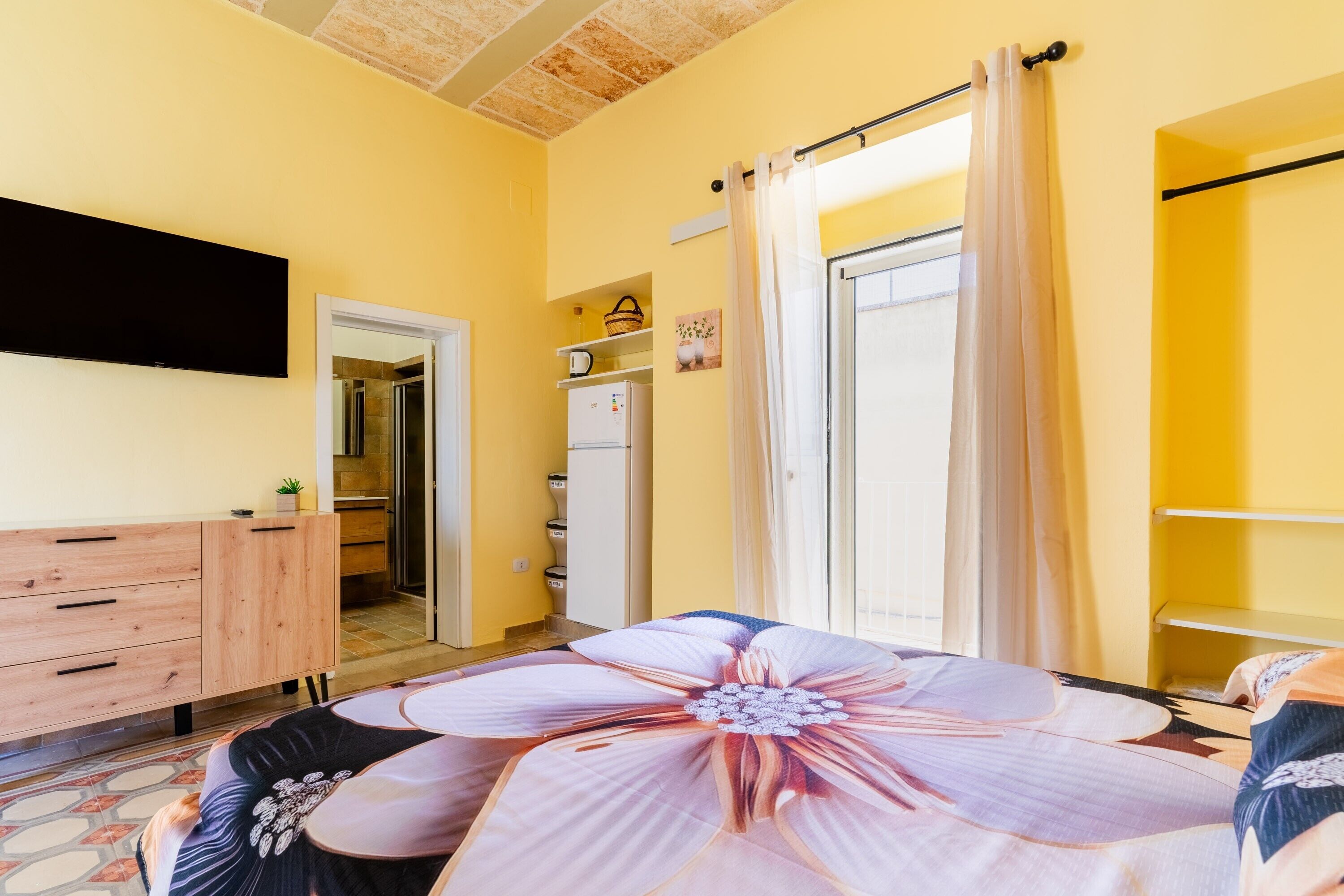 A bedroom with a large bed, a TV mounted on the wall, a wardrobe, and a door leading to a bathroom.
