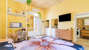 A bedroom with a large floral bedspread, a desk with a computer, a TV mounted on the wall, and a kitchen area visible through an open door.