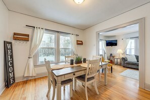 A dining area with a wooden table, white chairs, and a 'WELCOME' sign. There are windows with curtains, a TV, and a sofa in the background.