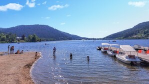 Une scène au bord du lac, avec des bateaux amarrés, des gens qui se baignent et une colline boisée en arrière-plan.