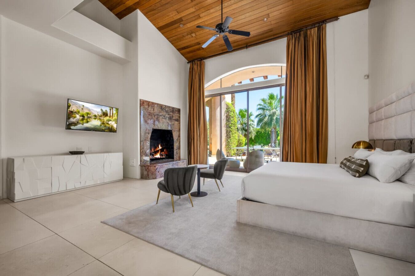 A modern hotel room with a fireplace, a large bed, a dining area, and a view of palm trees.