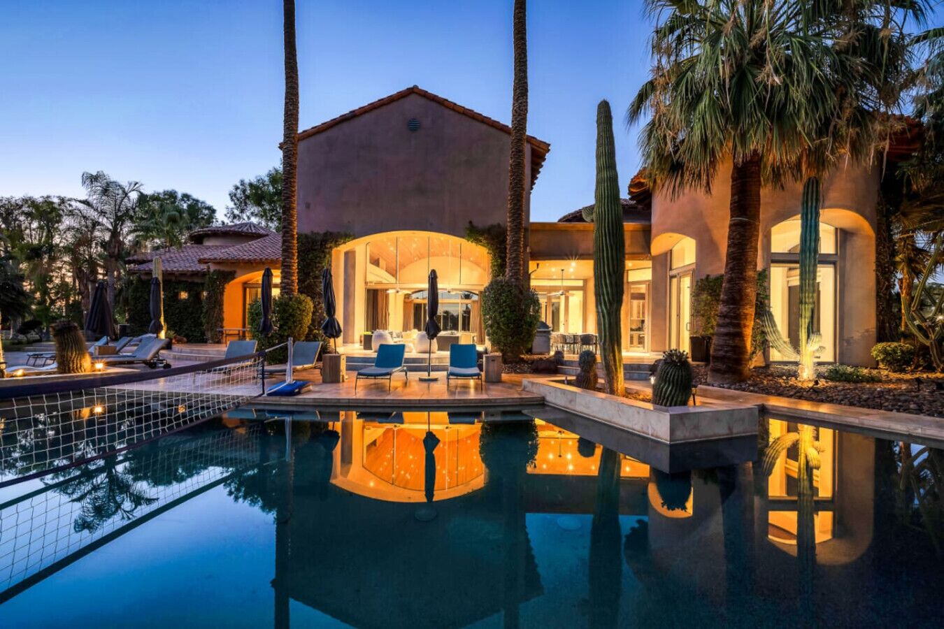 A pool with lounge chairs and umbrellas, a building with a red-tiled roof, and cacti in the foreground.