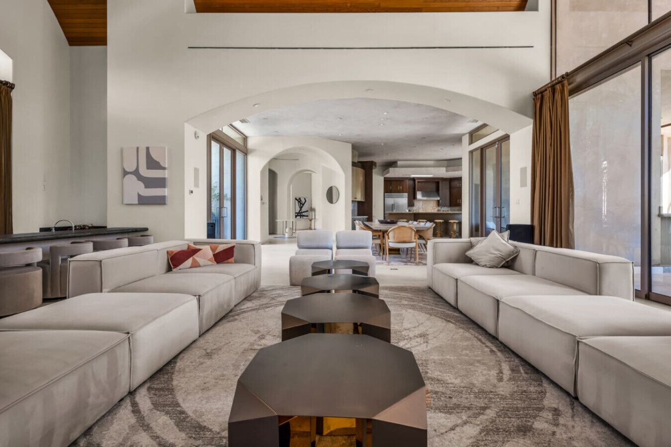 A spacious living room with a modern design, featuring a large sofa set, a coffee table, and a kitchen visible through an archway.