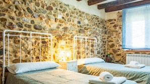 Una habitación con dos camas, paredes de piedra, una ventana con cortinas azules y una mesita de noche con lámpara.