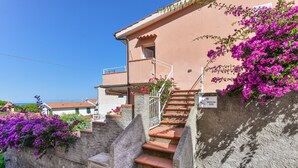 Ein Gebäude mit einer Treppe, die zu einer Tür mit der Bezeichnung 'ROOM 200' führt, umgeben von Steinmauern und leuchtend violetten Blumen.