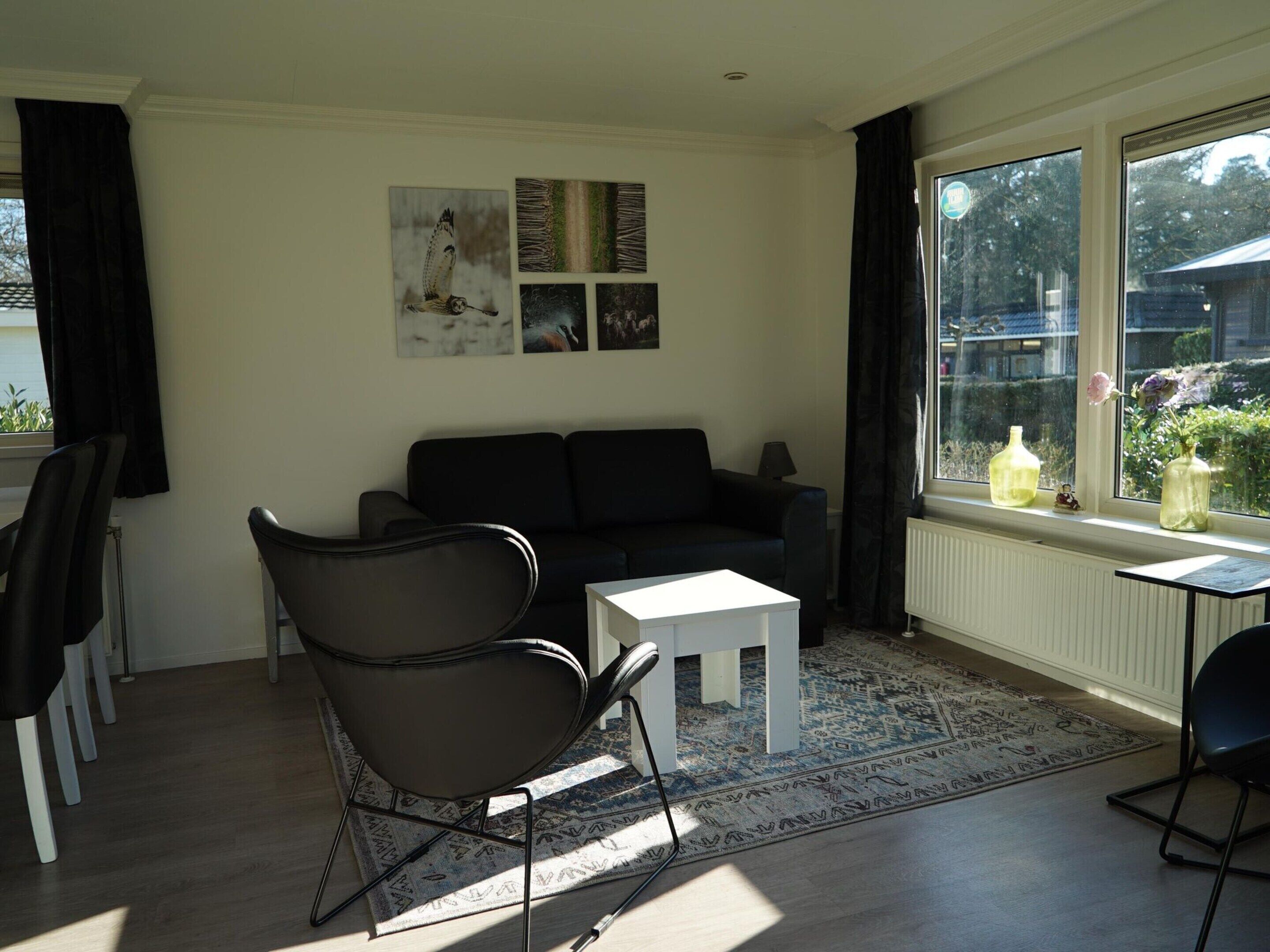 A living room with a black sofa, a white coffee table, and a patterned rug. There are two black chairs, a small white table, and a window with curtains.