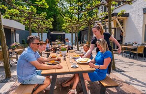 A group of people dining outdoors at a wooden table with a white building in the background. - Chalet in a Residence Near sea (Arnhem)