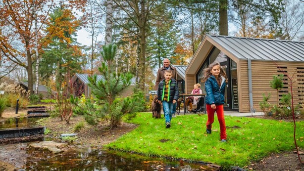 A family walking towards a modern cabin in a wooded area with a small pond. - Chalet in a Residence Near sea (Arnhem)